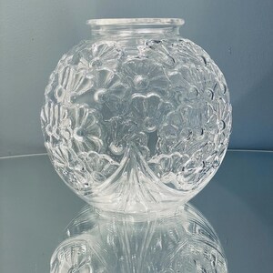 Könnte beinhalten: Eine klare, kugelförmige Glasvase mit floralem Muster. Die Vase hat eine weite Öffnung und steht auf einer reflektierenden Oberfläche, wodurch ein Spiegeleffekt entsteht. Das aufwendige Design zeigt detaillierte Blumen- und Blattmotive.
