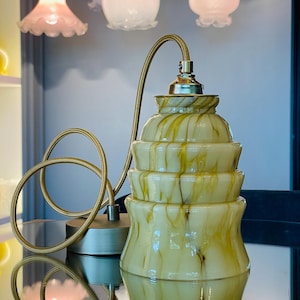 May include: A tiered glass pendant light fitting featuring a cream and olive green marbled swirl pattern. The lamp has a metallic gold-toned socket and a matching braided fabric cord that loops gracefully beside the glass shade.