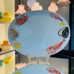 May include: An oval white ceramic serving platter featuring raised, three-dimensional decorative elements including a red crab, a red lobster, a brown scallop shell, a black mussel, and two yellow lemon slices arranged around the scalloped edge.