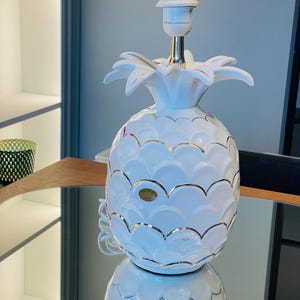 Puede incluir: Lámpara de mesa de cerámica blanca en forma de piña con detalles dorados. La lámpara tiene un cable blanco y una pantalla blanca.