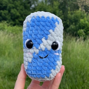 Può includere: Un giocattolo cilindrico di peluche con un volto sorridente, grandi occhi neri e guance rosa. La superficie del giocattolo è un mix di blu e grigio, che ricorda un globo. Lo sfondo è una vista sfocata di erba verde e alberi.