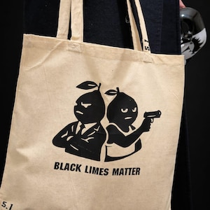 May include: Beige tote bag featuring a black graphic of two lime-shaped figures, one in a suit and the other holding a gun. The text "BLACK LIMES MATTER" is printed on the bag. It has two beige handles.