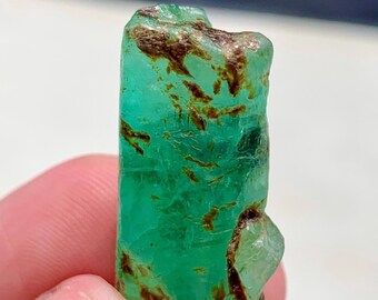 Puede incluir: Una esmeralda en bruto sin cortar. La gema es de color verde vibrante con inclusiones marrones y una textura natural rugosa. La esmeralda se sostiene entre los dedos, mostrando su forma y color naturales.