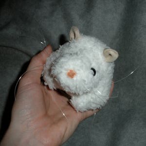 Op de afbeelding: Wit pluche hamster speelgoed met lange witte snorharen. Het speelgoed heeft een roze neus en zwarte ogen.