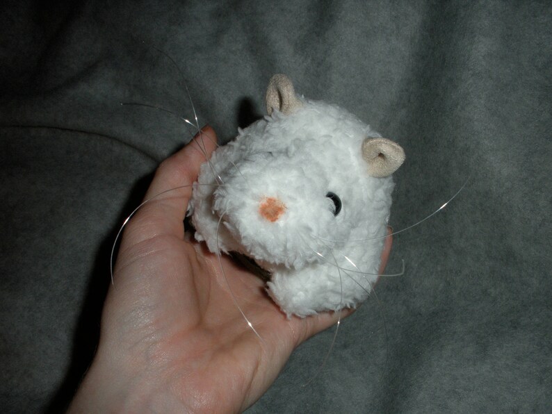 Op de afbeelding: Wit pluche hamster speelgoed met lange witte snorharen. Het speelgoed heeft een roze neus en zwarte ogen.