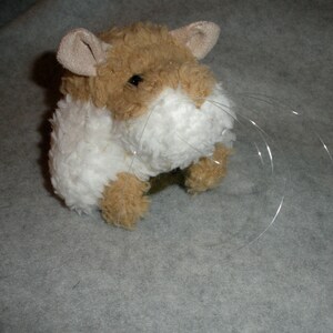 Op de afbeelding: Een bruin en wit pluche hamster speelgoed met lange witte snorharen. Het speelgoed zit op een grijs oppervlak.