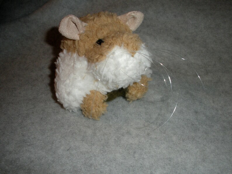 Op de afbeelding: Een bruin en wit pluche hamster speelgoed met lange witte snorharen. Het speelgoed zit op een grijs oppervlak.