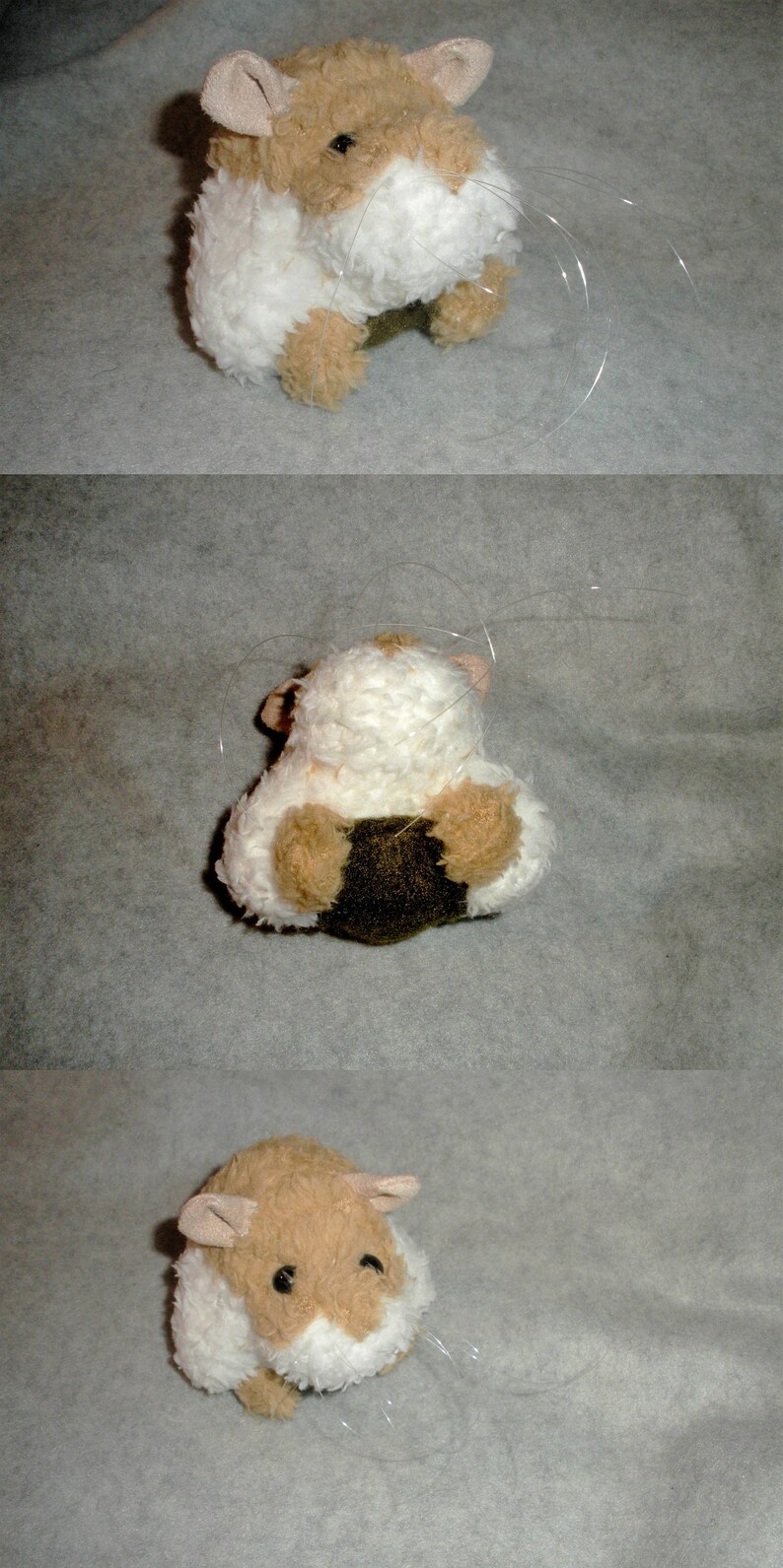 Op de afbeelding: Een bruin en wit pluche hamster speelgoed met lange witte snorharen. Het speelgoed zit op een lichtgrijze ondergrond.