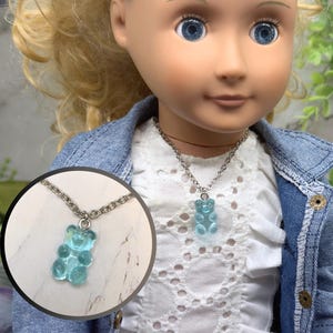 May include: A light blue gummy bear pendant necklace. The pendant is a translucent blue color and hangs from a silver-colored chain. The necklace is displayed on a doll wearing a denim jacket and white shirt.