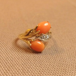 May include: A gold ring featuring two oval, orange-red gemstones. The ring has a leaf-like design with a small section of white metal with small, clear stones. The ring is set against a neutral background.