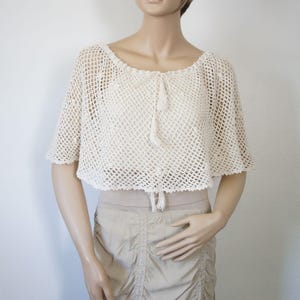 May include: A cream-colored, short-sleeved crochet top featuring an open-knit diamond pattern, a wide scoop neckline, and a central drawstring closure with two hanging tassels. The garment has a cropped length and a scalloped edge finish.