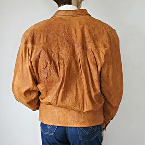 K&ouml;nnte beinhalten: Eine braune Lederjacke mit Knopfleiste und langen &Auml;rmeln. Die Jacke hat eine knitterige Textur und eine leicht &uuml;berdimensionierte Passform.