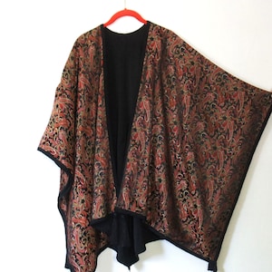 May include: A black open-front poncho featuring a detailed paisley pattern in shades of red, gold, and dark green. The garment has a solid black interior lining, a wide draped silhouette, and black trim along the edges with small tassels at the hem.