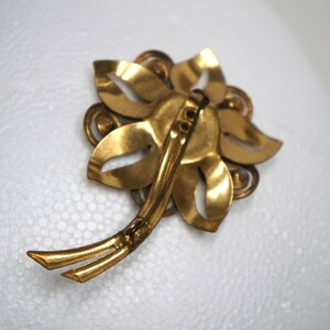 May include: Gold-toned metal flower brooch with a pin closure on the back. The brooch is made of several overlapping leaves with a decorative center.