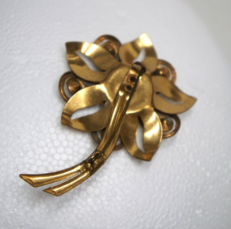 May include: Gold-toned metal flower brooch with a pin closure on the back. The brooch is made of several overlapping leaves with a decorative center.