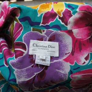 K&ouml;nnte beinhalten: Ein wei&szlig;es Etikett mit schwarzem Text, auf dem "Christian Dior Separates Paris New York" und Gr&ouml;&szlig;eninformationen stehen. Das Etikett befindet sich auf einem Stoffhintergrund mit einem floralen Muster in den Farben Rosa, Lila, Gelb und T&uuml;rkis.