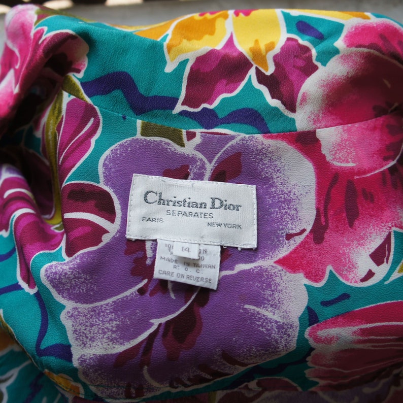 K&ouml;nnte beinhalten: Ein wei&szlig;es Etikett mit schwarzem Text, auf dem "Christian Dior Separates Paris New York" und Gr&ouml;&szlig;eninformationen stehen. Das Etikett befindet sich auf einem Stoffhintergrund mit einem floralen Muster in den Farben Rosa, Lila, Gelb und T&uuml;rkis.