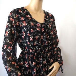 K&ouml;nnte beinhalten: Ein schwarzes, lang&auml;rmliges Kleid mit einem Blumendruck in Rosa-, Rot- und Wei&szlig;t&ouml;nen. Das Kleid hat einen V-Ausschnitt und eine Schleife am Ausschnitt.