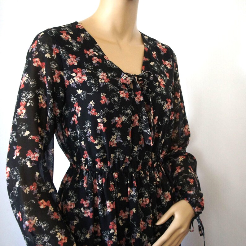 K&ouml;nnte beinhalten: Ein schwarzes, lang&auml;rmliges Kleid mit einem Blumendruck in Rosa-, Rot- und Wei&szlig;t&ouml;nen. Das Kleid hat einen V-Ausschnitt und eine Schleife am Ausschnitt.