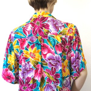 K&ouml;nnte beinhalten: Ein kurz&auml;rmeliges Hemd mit einem leuchtenden Blumendruck. Das Hemd hat einen t&uuml;rkisfarbenen Hintergrund mit rosa, gelben, violetten und gr&uuml;nen Blumen.
