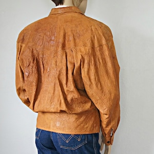 K&ouml;nnte beinhalten: Eine braune Lederjacke mit Knopfleiste und langen &Auml;rmeln. Die Jacke hat eine strukturierte, knitterige Optik.
