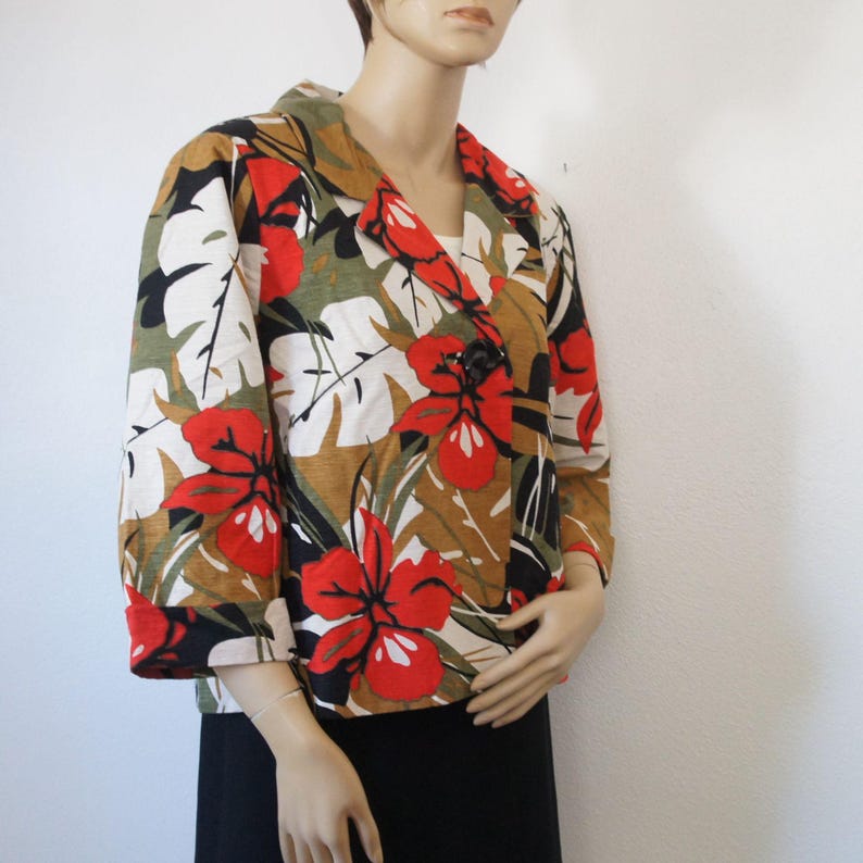 Puede incluir: Una blazer de manga tres cuartos con un estampado floral vibrante. La chaqueta presenta grandes flores de hibisco rojas, hojas blancas y follaje verde sobre un fondo marr&oacute;n y negro. Tiene un cierre de bot&oacute;n negro &uacute;nico y un cuello con muescas.