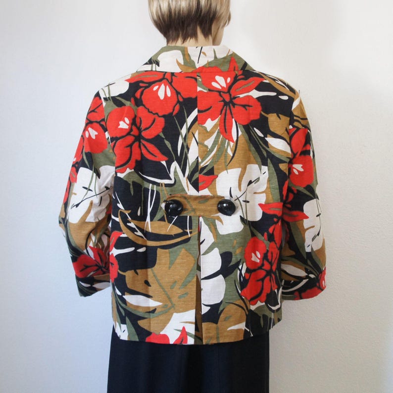 Puede incluir: Chaqueta corta con un llamativo estampado floral. La chaqueta presenta grandes flores rojas, hojas blancas y detalles negros sobre un fondo beige y verde oliva. Tiene dos grandes botones negros y un cuello.