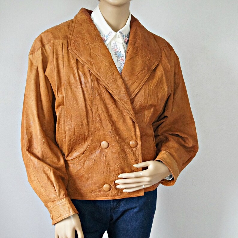 K&ouml;nnte beinhalten: Eine braune Lederjacke mit doppelreihigem Verschluss und zwei gro&szlig;en Kn&ouml;pfen. Die Jacke hat einen breiten Kragen und eine leicht verk&uuml;rzte L&auml;nge.