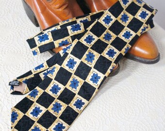 May include: A black and gold patterned silk tie with blue floral accents. The tie is folded and placed on top of a pair of brown cowboy boots.
