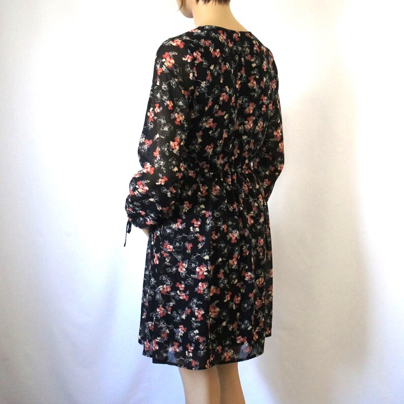 K&ouml;nnte beinhalten: Ein schwarzes Blumenkleid mit langen &Auml;rmeln und einer taillierten Taille. Das Kleid hat einen kleinen Blumendruck in Rosa- und Wei&szlig;t&ouml;nen.