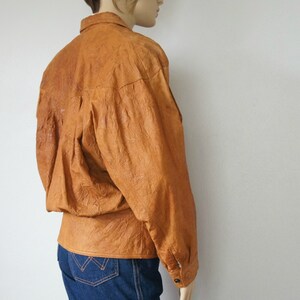 K&ouml;nnte beinhalten: Eine braune Lederjacke mit Knopfleiste und langen &Auml;rmeln. Die Jacke hat eine strukturierte, knitterige Optik.