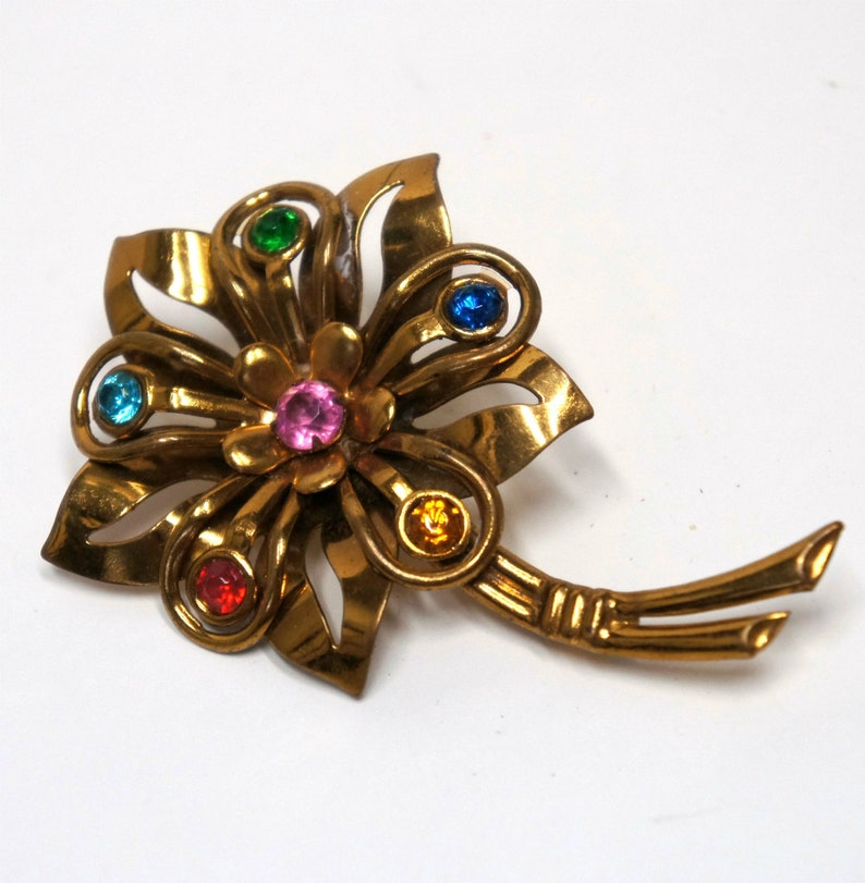 May include: A gold-toned flower brooch with five colorful rhinestones. The brooch has a delicate, intricate design and a long, thin stem.