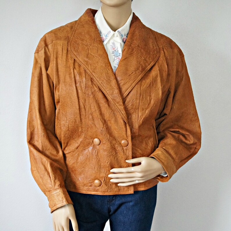 K&ouml;nnte beinhalten: Eine braune Lederjacke mit doppelreihigem Verschluss und einem gro&szlig;en Kragen. Die Jacke hat eine strukturierte Oberfl&auml;che und zwei Kn&ouml;pfe auf der Vorderseite.