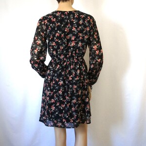K&ouml;nnte beinhalten: Ein schwarzes Blumenkleid mit langen &Auml;rmeln und einem V-Ausschnitt. Das Kleid hat einen figurbetonten Oberteil und einen weiten Rock.
