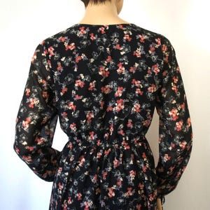K&ouml;nnte beinhalten: Ein schwarzes, lang&auml;rmliges Kleid mit einem Blumendruck in Rosa-, Rot- und Wei&szlig;t&ouml;nen. Das Kleid hat eine eingezogene Taille und einen V-Ausschnitt.