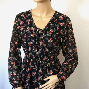 K&ouml;nnte beinhalten: Ein schwarzes, lang&auml;rmliges Kleid mit floralem Muster und einem Knoten am Ausschnitt. Das Kleid hat einen taillierten Oberteil und einen flie&szlig;enden Rock.