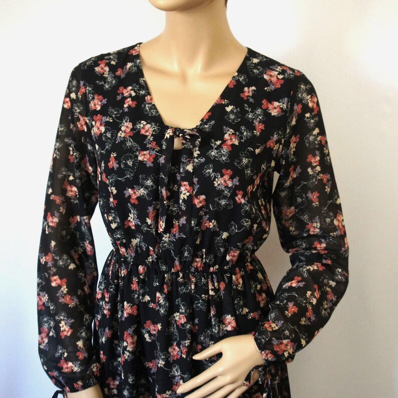 K&ouml;nnte beinhalten: Ein schwarzes, lang&auml;rmliges Kleid mit floralem Muster und einem Knoten am Ausschnitt. Das Kleid hat einen taillierten Oberteil und einen flie&szlig;enden Rock.