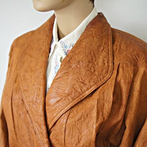 K&ouml;nnte beinhalten: Eine braune Lederjacke mit strukturierter Oberfl&auml;che und einem gro&szlig;en Kragen. Die Jacke ist offen und hat darunter ein wei&szlig;es Hemd mit floralem Muster.