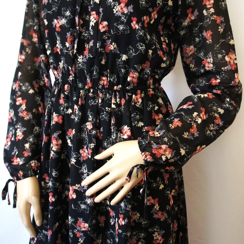 K&ouml;nnte beinhalten: Ein schwarzes, lang&auml;rmliges Kleid mit floralem Muster, einer gerafften Taille und Bindedetails an den &Auml;rmeln.