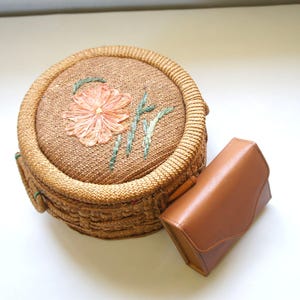 May include: A round, woven straw bag featuring a light orange embroidered flower and green stems on the lid, paired with a small, rectangular, smooth tan leather clutch with a flap closure and a curved front edge.
