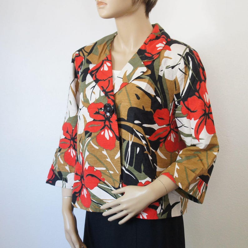 Puede incluir: Una blazer de manga corta con un estampado floral llamativo. La chaqueta presenta grandes flores rojas, hojas verdes y detalles negros sobre un fondo beige y blanco. Tiene un cierre de bot&oacute;n negro &uacute;nico y un dise&ntilde;o de solapa cl&aacute;sico.