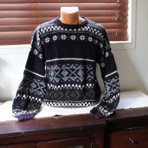 Könnte beinhalten: Ein langärmeliger Pullover mit Rundhalsausschnitt und einem geometrischen Muster in Dunkellila, Schwarz und Weiß. Das Strickkleidungsstück zeigt horizontale Bänder aus Rauten, Quadraten und komplexen Formen auf Brust, Ärmeln und Saum.