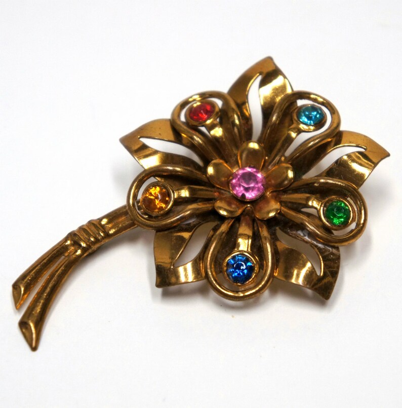 May include: A gold-toned flower brooch with a stem and five colorful rhinestones. The rhinestones are red, orange, pink, blue, and green.