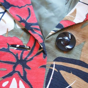 Puede incluir: Primer plano de una blazer con un vibrante estampado floral en rojo, azul marino y blanco sobre un fondo caqui. La blazer presenta un gran bot&oacute;n negro y una solapa con muescas. La tela tiene un aspecto texturizado.