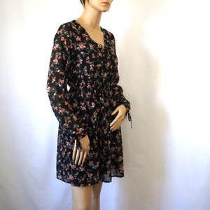 K&ouml;nnte beinhalten: Ein schwarzes Kleid mit floralem Muster, langen &Auml;rmeln und einem Knoten am Hals. Das Kleid hat einen figurbetonten Oberteil und einen weiten Rock.