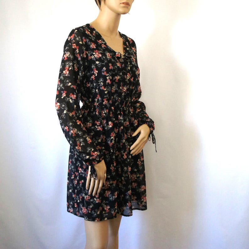 K&ouml;nnte beinhalten: Ein schwarzes Kleid mit floralem Muster, langen &Auml;rmeln und einem Knoten am Hals. Das Kleid hat einen figurbetonten Oberteil und einen weiten Rock.