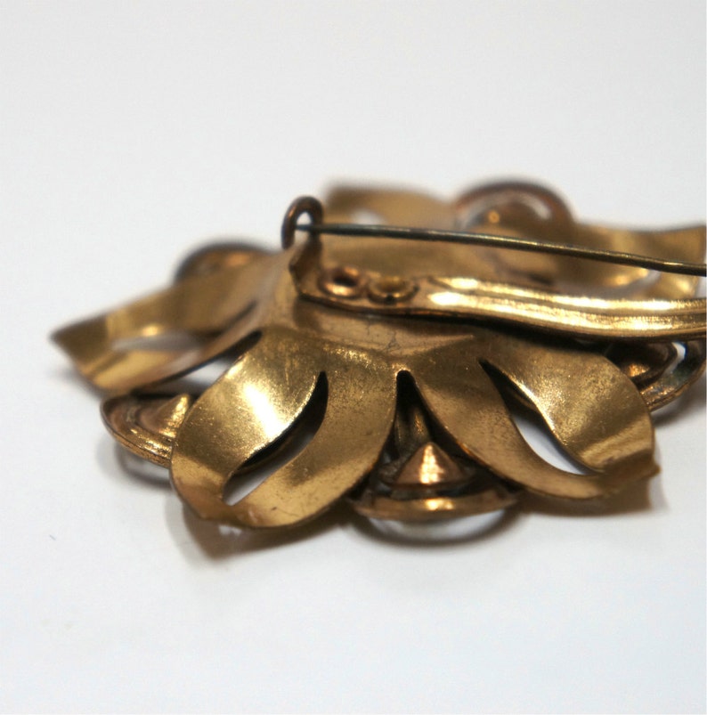 May include: A gold-toned brooch with a flower design and clear rhinestones. The brooch has a pin back.