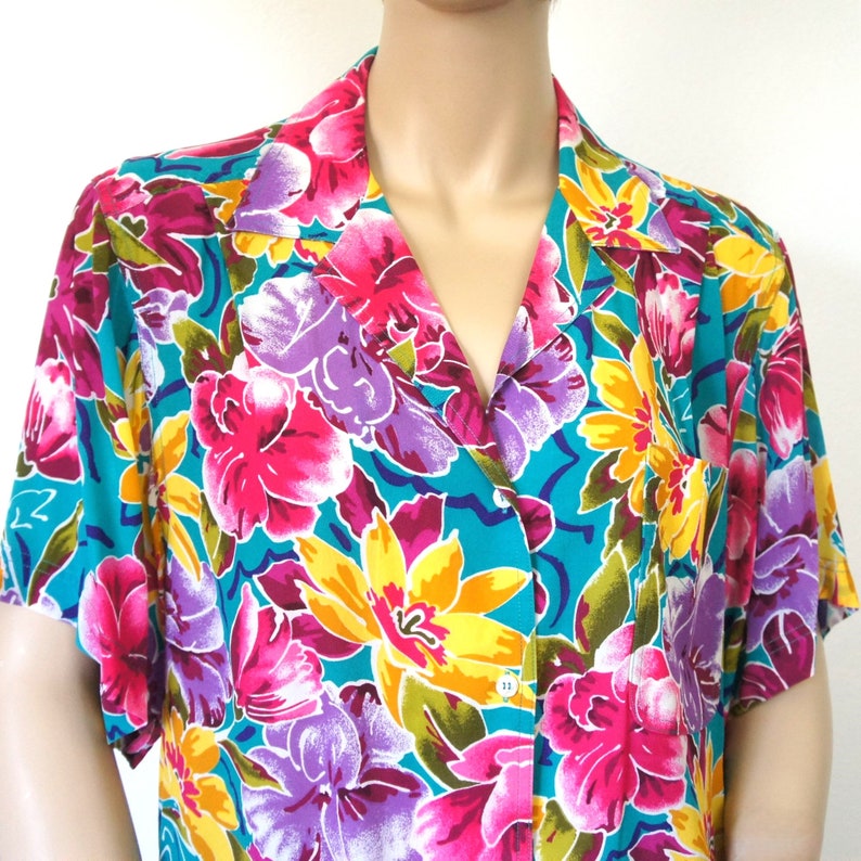 K&ouml;nnte beinhalten: Ein t&uuml;rkisfarbenes Kurzarmhemd mit einem Blumendruck. Der Druck zeigt gro&szlig;e rosa, gelbe und lila Blumen auf einem t&uuml;rkisfarbenen Hintergrund.