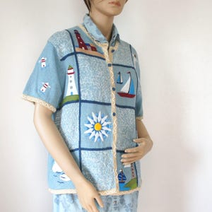 May include: Light blue short-sleeved button-up shirt with nautical-themed embroidered patches. The patches feature lighthouses, sailboats, and a sun design. The shirt has a light beige trim and a collared neckline.