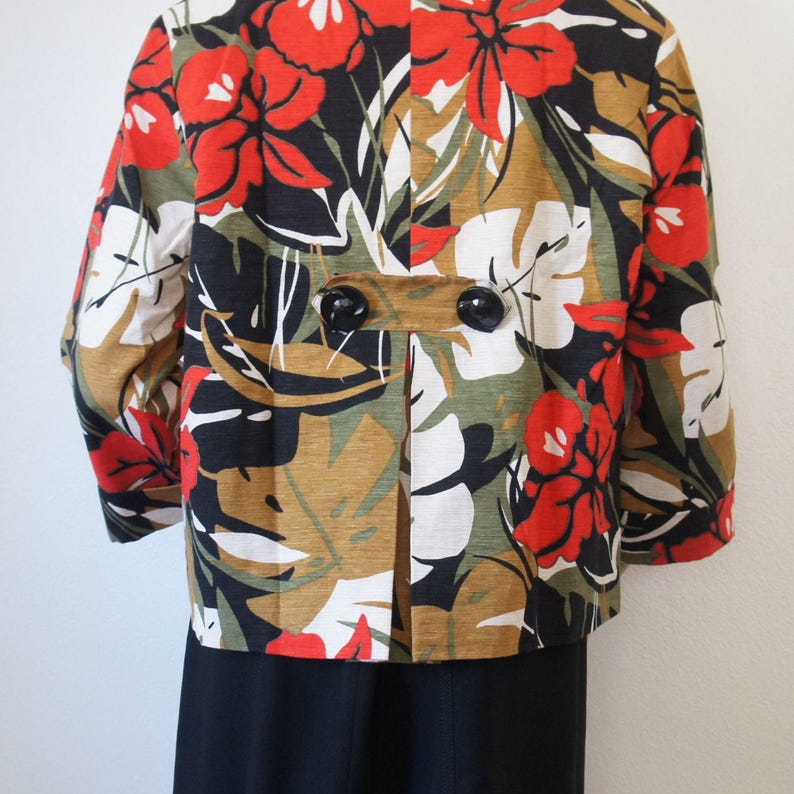 Puede incluir: Chaqueta corta con un vibrante estampado floral con flores y hojas rojas, blancas, negras y verde oliva. La chaqueta tiene grandes botones negros y mangas tres cuartos. La parte inferior de la chaqueta es negra.